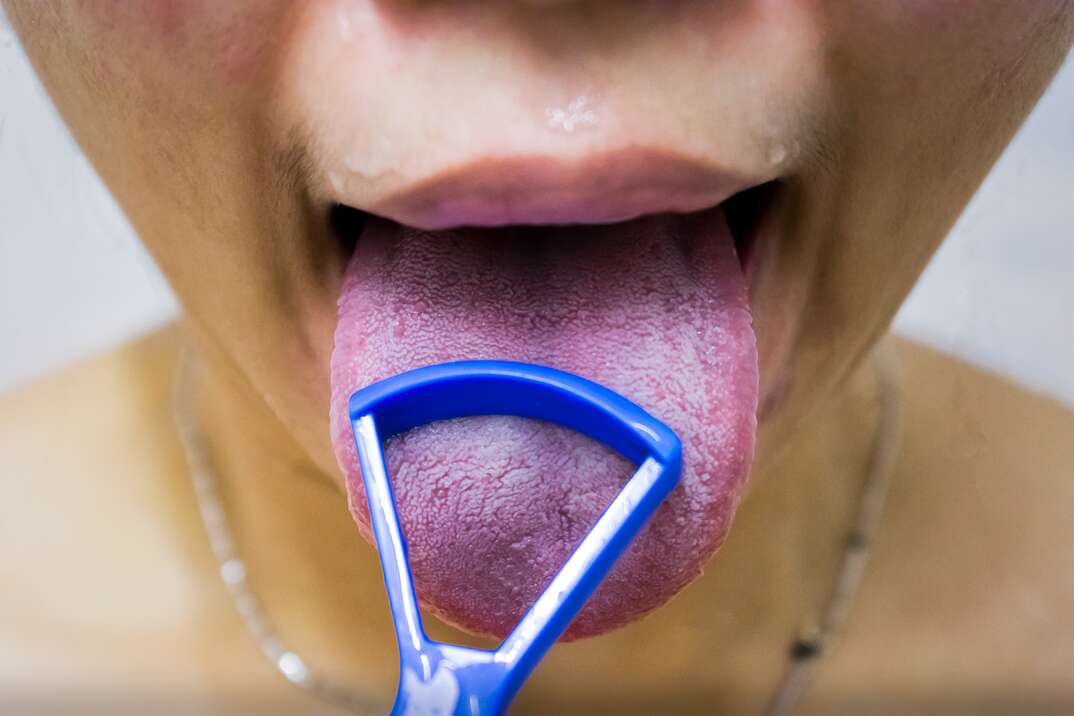 An extreme closeup of the lower face of what appears to be a woman with her mouth open and tongue sticking out uses a tongue scraper oral hygiene instrument, tongue scraper, tongue, human tongue, oral hygiene, dental hygiene, dental, oral, medical, hygiene, woman, pink tongue, scraper, dental instrument