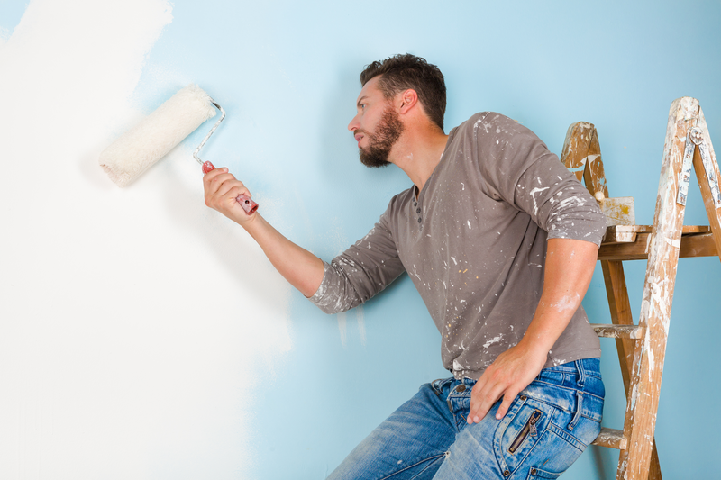 Portrait of handsome young painter in paint splattered shirt painting a wall with paint roller and leaning on a wooden ladder
