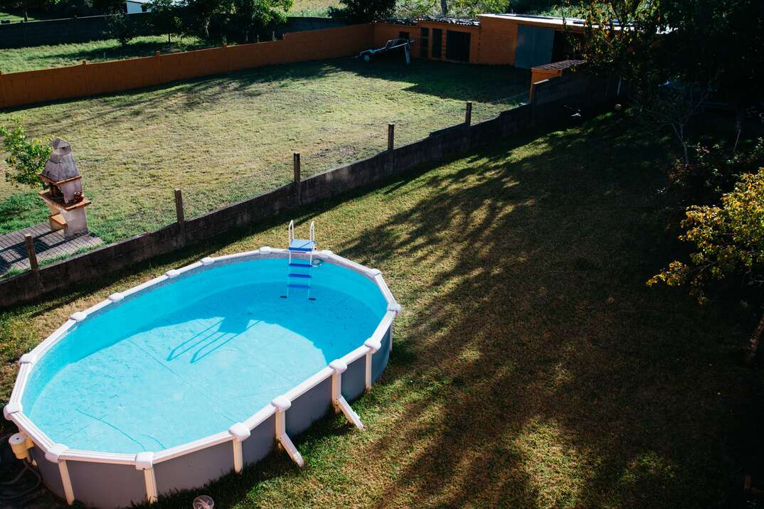 An above ground pool with bright blue water sits in a large and empty backyard 