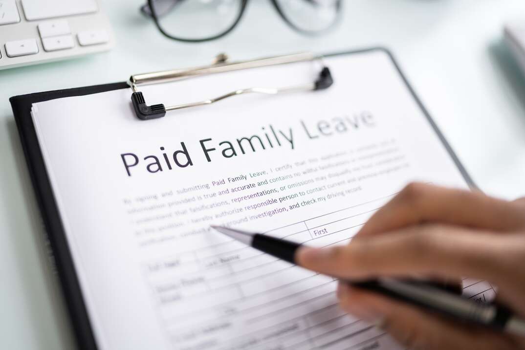 A male hand holding a pen points to a document on a clipboard resting on a white surface that reads Paid Family Leave in relation to the FMLA or Family Medical Leave Act as a pair of eyeglasses sits nearby, eyeglasses, glasses, clipboard, document, legal document, FMLA, Family Medical Leave Act, paid family leave, medical, medical document, healthcare, health care, pen, ink pen, human hand, hand, male, male hand