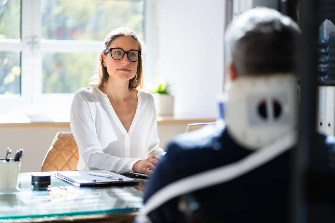 A man wearing a neck brace sits across a desk from a female personal injury attorney in a law office, man, male, injured man, injured male, neck brace, neck injury, injury, hurt, woman, female, lawyer, attorney, female lawyer, female attorney, law office, office, desk, personal injury, injury