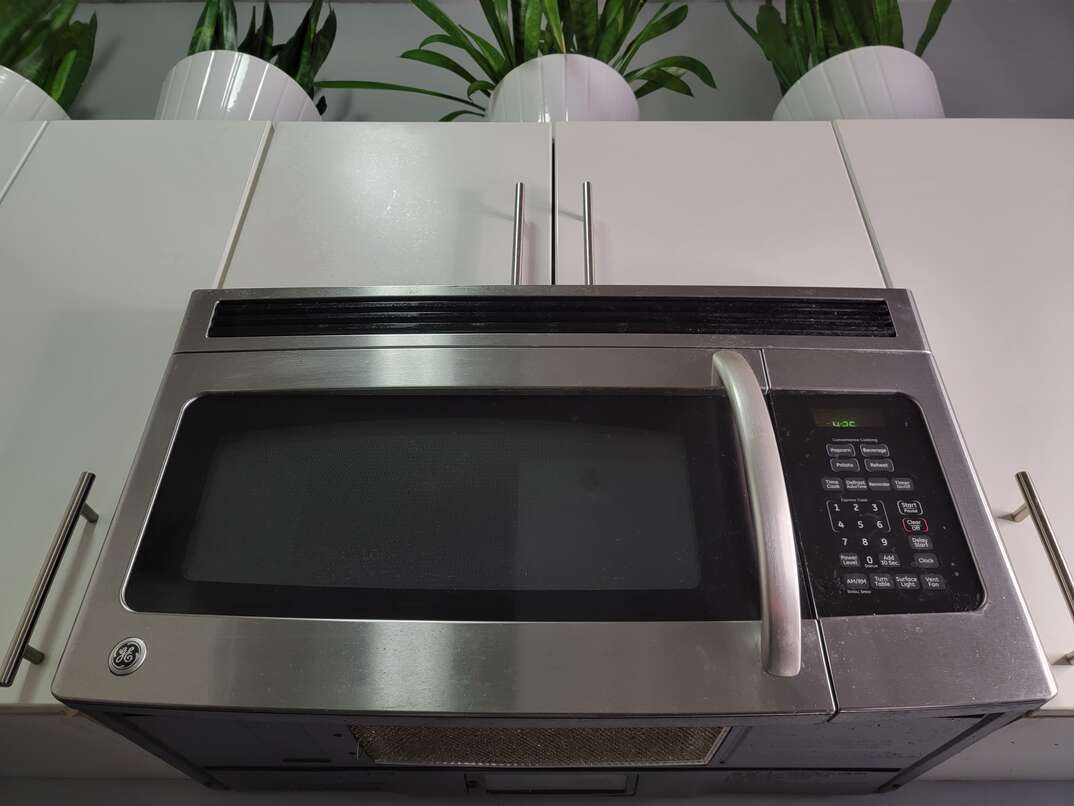 A stainless steel microwave mounted over the range amid white cabinetry and green plants on top of the cabinets