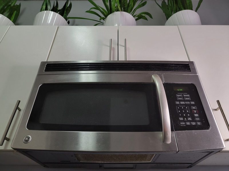 A stainless steel microwave mounted over the range amid white cabinetry and green plants on top of the cabinets, microwave, stainless steel microwave, stainless steel, steel, metal, metallic, black and silver, white cabinetry, cabinetry, cabinets, white cabinets, plants, green plants in white ceramic pots, white ceramic pots, ceramic pots, pots, green plants, plants