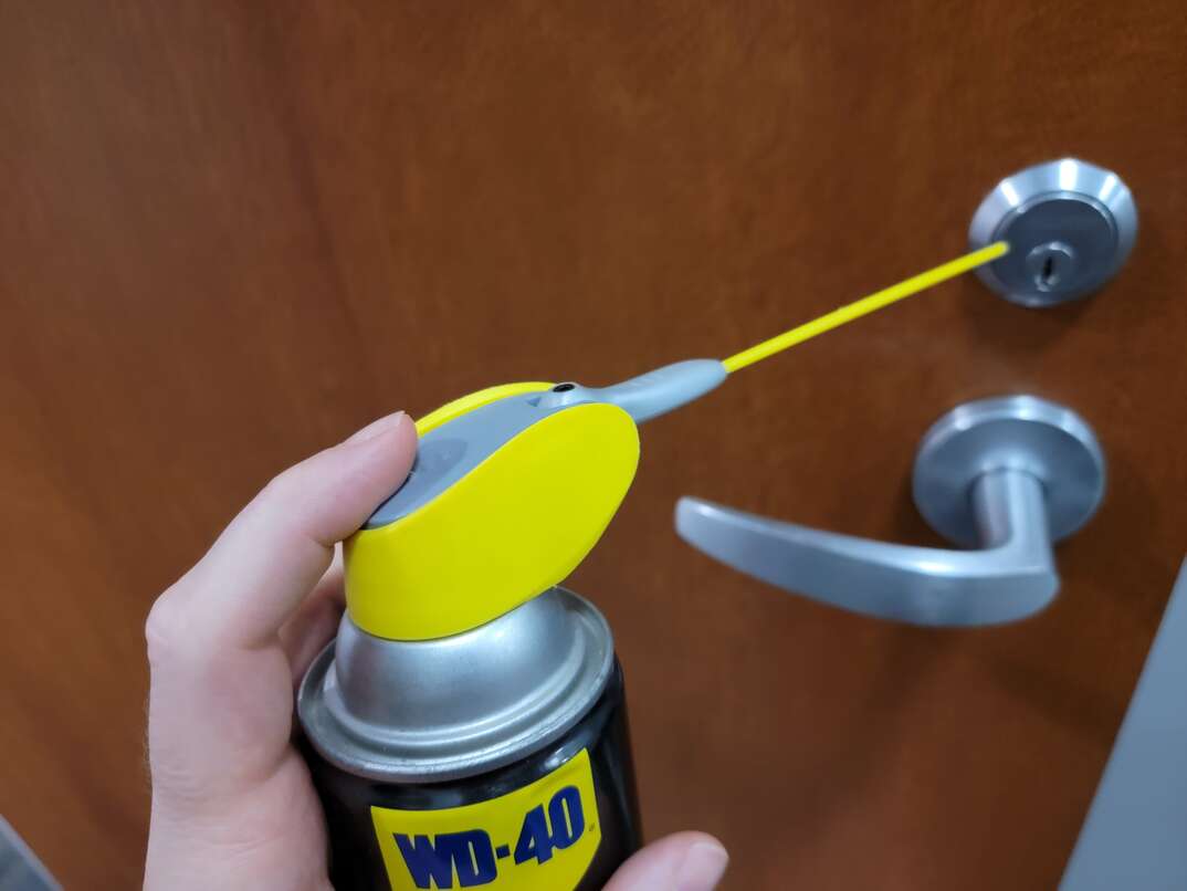 A male hand points the spray nozzle of a can of WD-40 at the lock of a front door, spray nozzle, nozzle, spray, sprayer, spraying, WD-40, spray can, hand, human hand, door, front door, residential door, door handle, door knob, doorknob, door lock, bolt lock, lubricant, lube, libricate