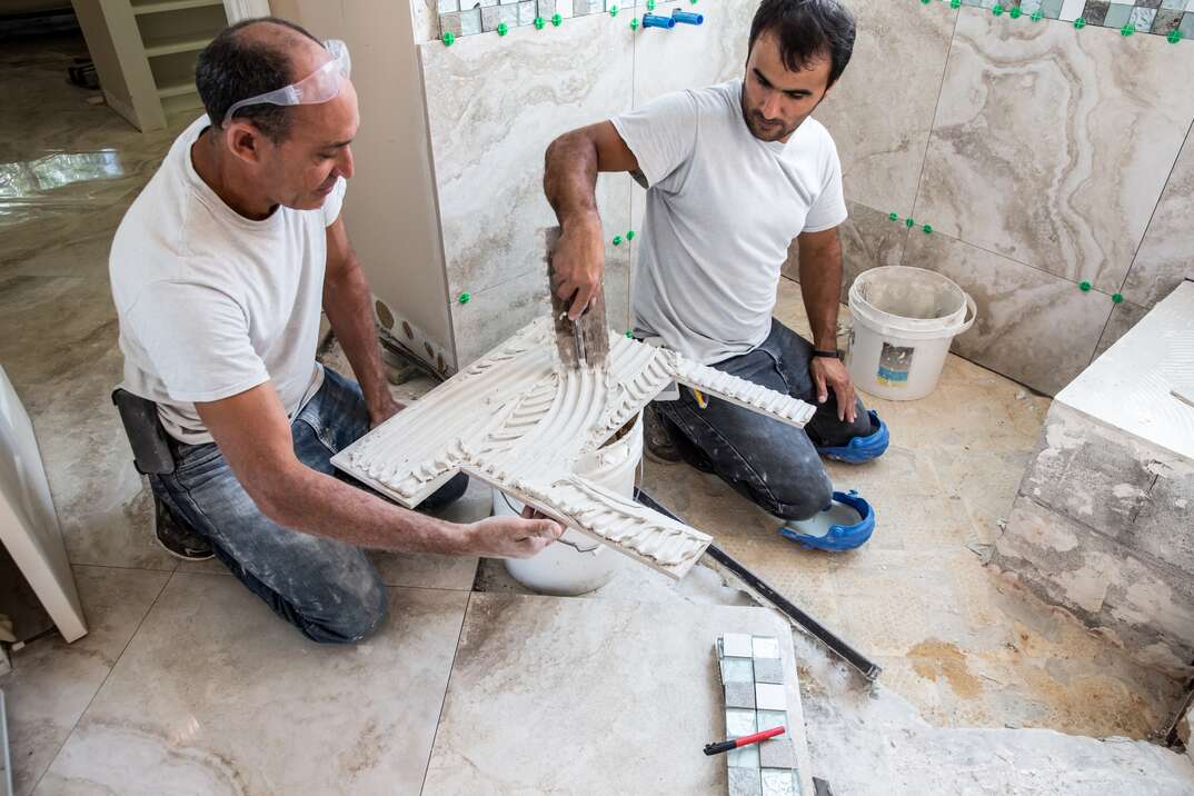 New tile is being installed in shower of a residential home that is being renovated  Two male tile installers are putting cement adhesive on the back of a natural stone tile that will be placed on the wall of the shower 