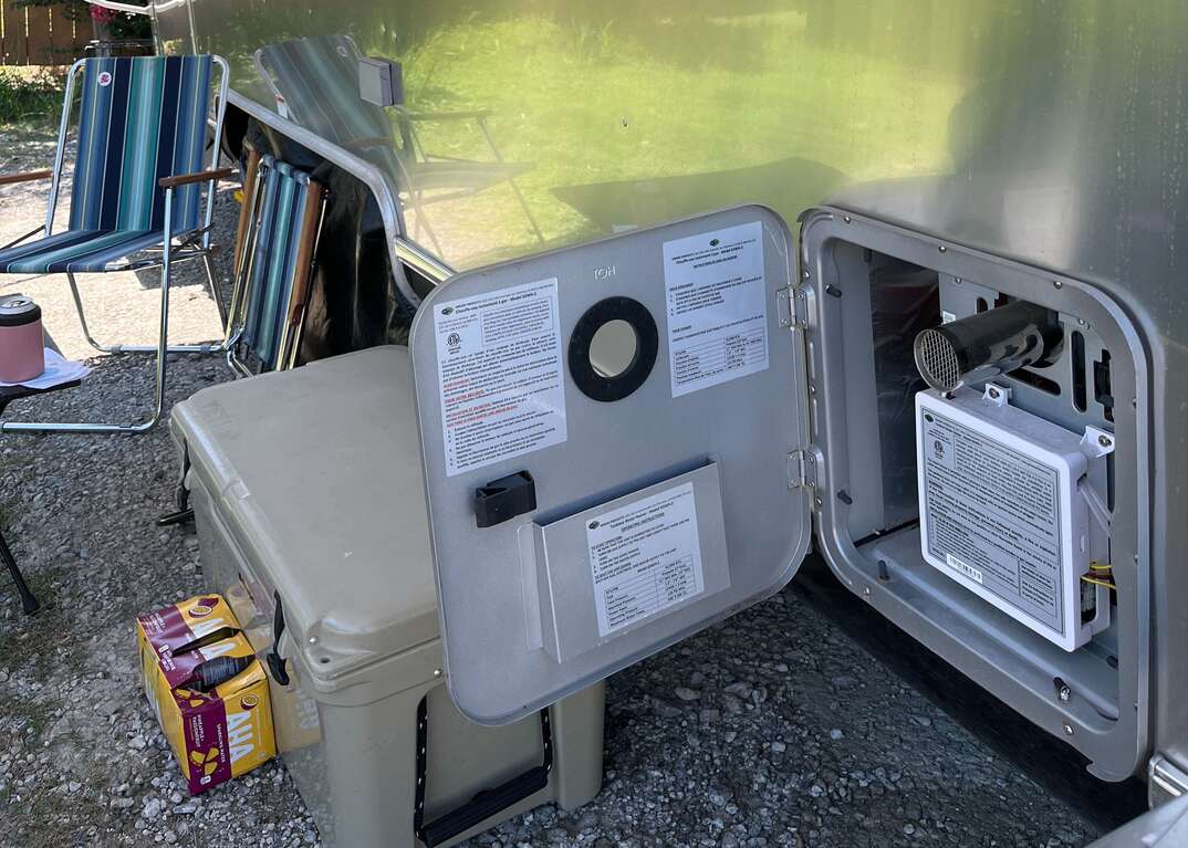 The door to the water heater is open on a stainless steel Airstream mobile home parked at a campsite with its retractable awning extended