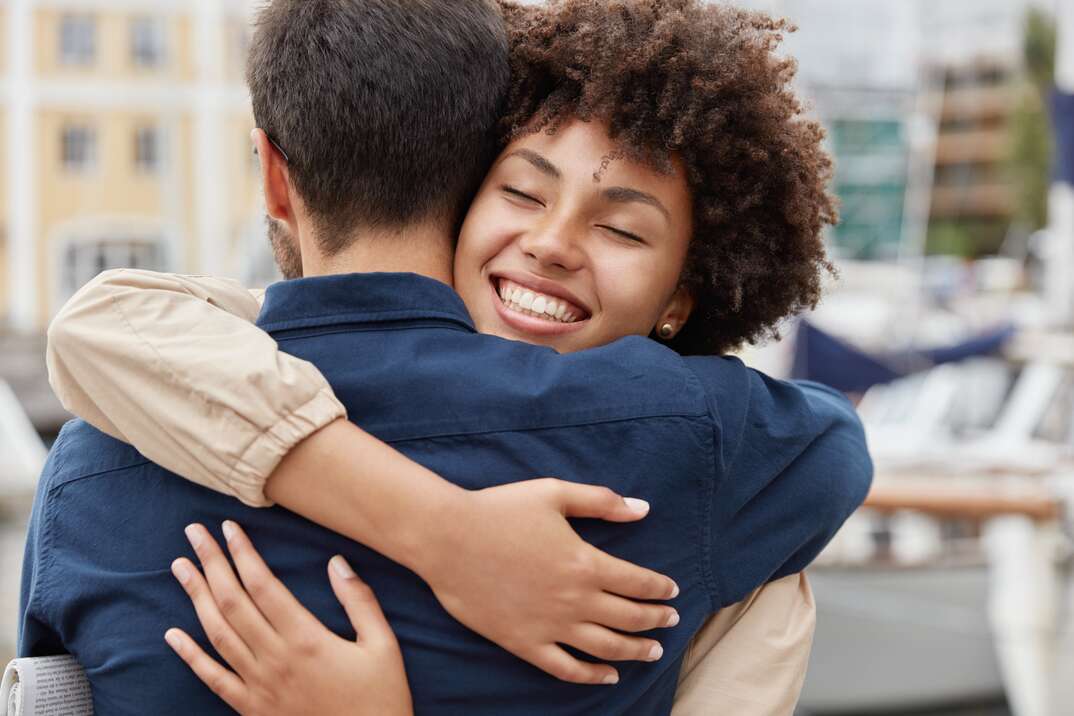 Farewell concept. Delighted happy smiling African American woman says goodbye to boyfriend who sails for long time, gives warm hug, pose together against harbour background. Truthful feelings