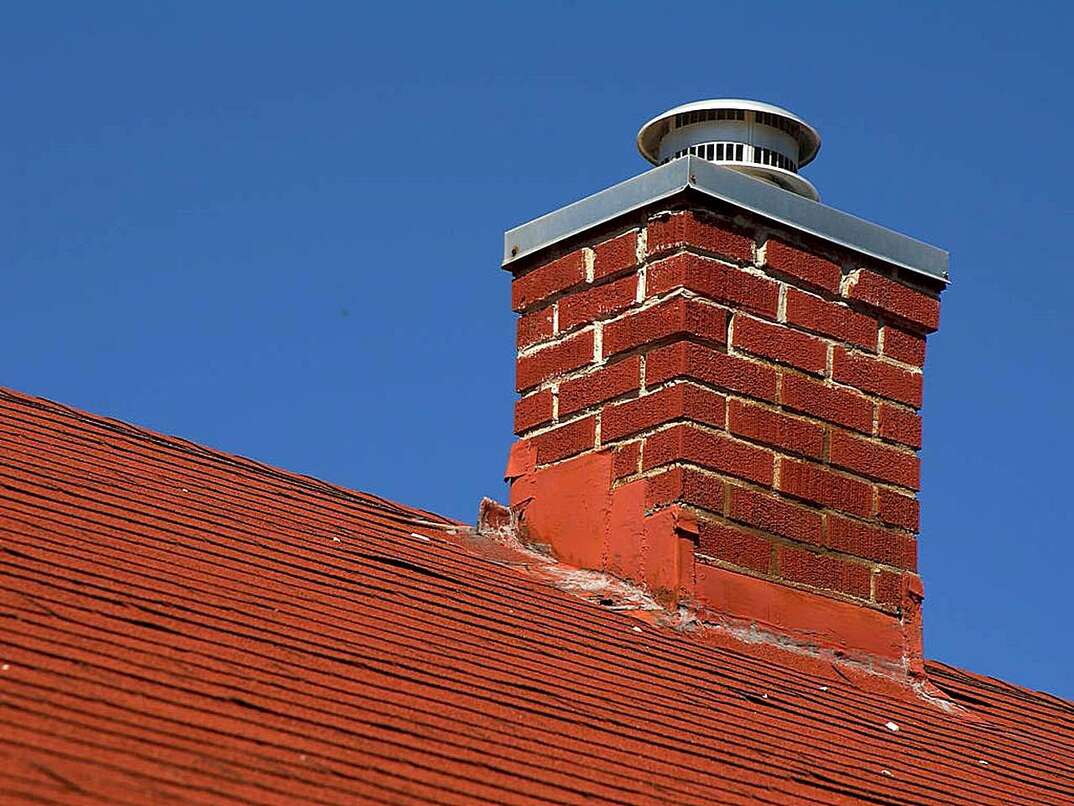 A red brick chimney in relative disrepair is seen rising from the red shingled roof of a house against a blue sky, chimney, brick chimney, red brick chimney, red shingled roof, shingled roof, roof, shingles, blue sky, sky, blue