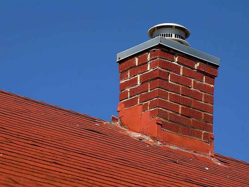 A red brick chimney in relative disrepair is seen rising from the red shingled roof of a house against a blue sky, chimney, brick chimney, red brick chimney, red shingled roof, shingled roof, roof, shingles, blue sky, sky, blue