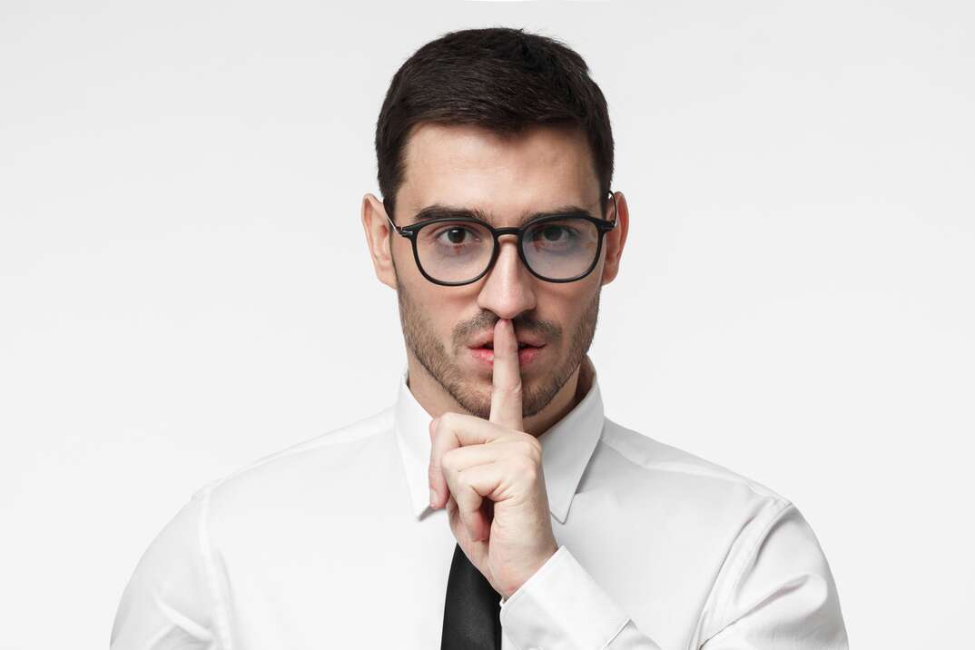 A male business professional in a white shirt and black tie holds his finger over his lips in a secretive gesture as he stands against a white background, white background, male professional, professional, male, white shirt, black tie, eyeglasses, shh, be quiet, secretive, secretive gesture, finger over lips, finger, looking into camera