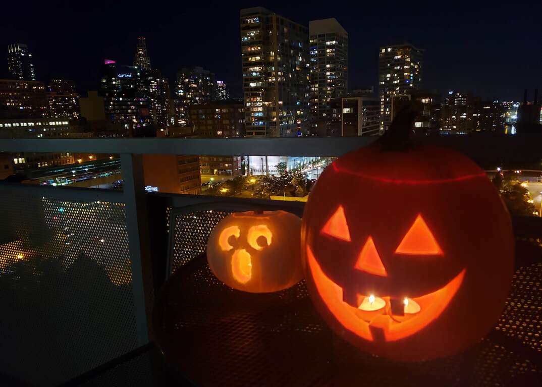 Two glowing, a large orange one and a small white one, candlelit Halloween jack-o-lanterns, sit on a patio table on the balcony of a high rise residence with an urban skyline in the background.