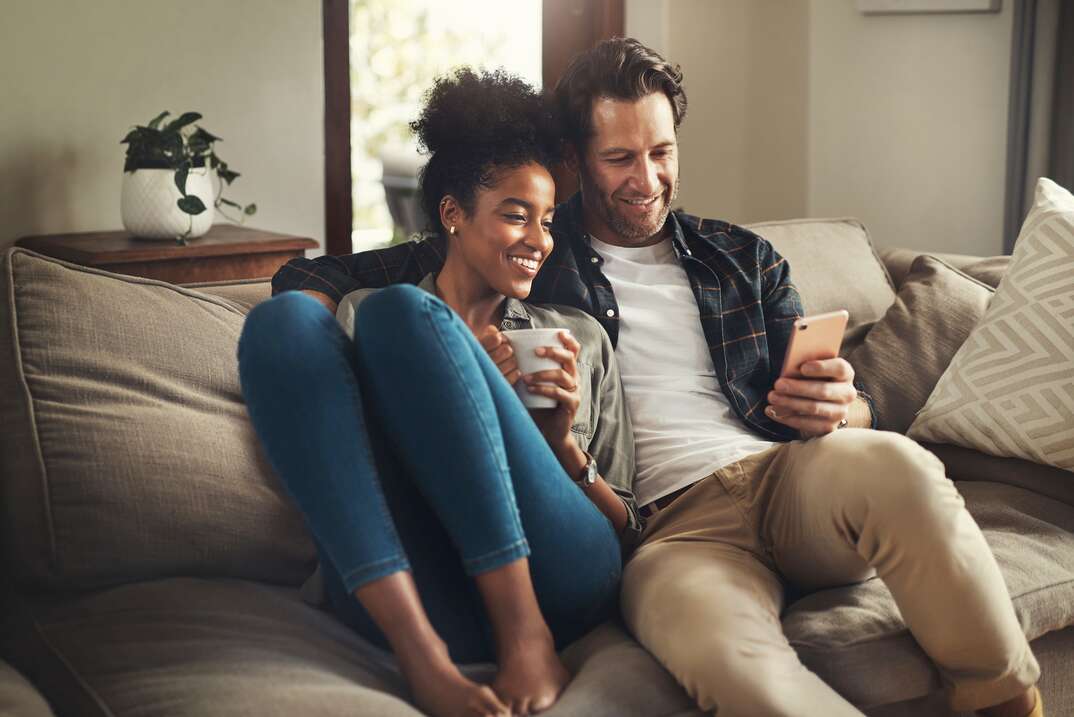 A male and female married couple sit close together on a couch in the living room and appear amused while looking at the smartphone the man is holding, male and female, male, female, man, woman, male and female couple, married couple, couple, married, husband, wife, husband and wife, sitting on couch, couch, living room, love, close, sitting close, looking at phone, smartphone, living room, house, home, living room