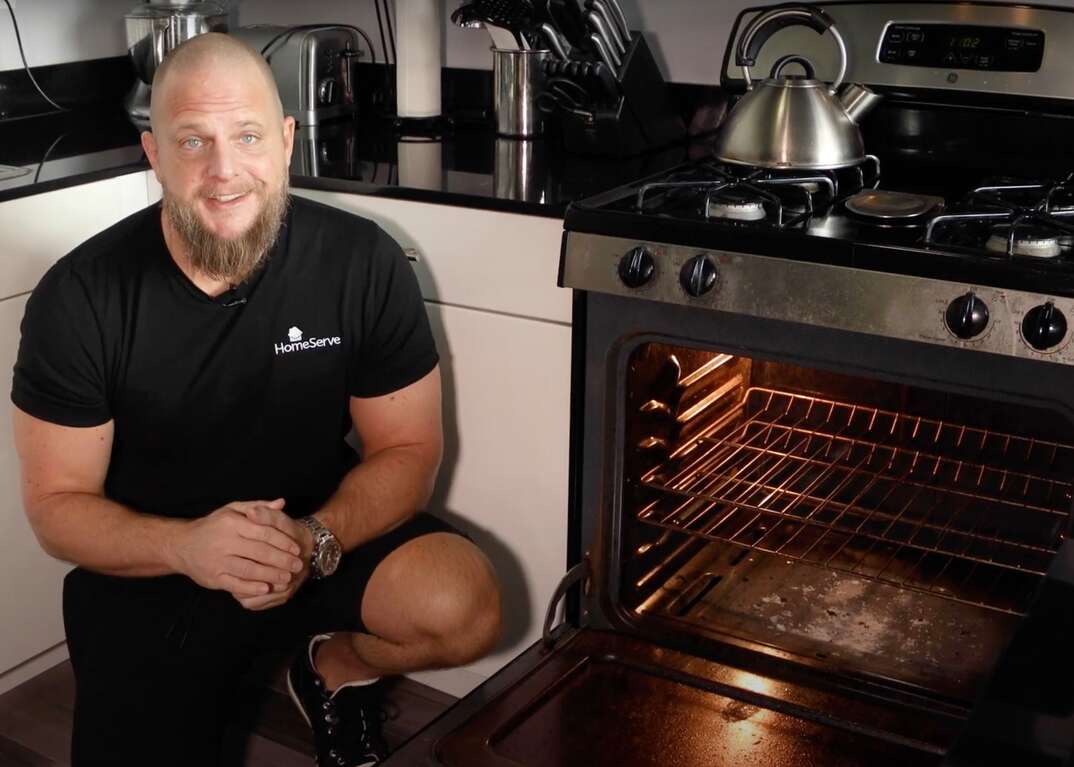 HomeServe Managing Editor Matt Schmitz crouches in a kitchen next to an open oven that clearly has not been cleaned in a very long time, dirty oven, oven cleaning, oven, cleaning, maintenance, appliance maintenance, kitchen appliance, appliance, male, Matt Schmitz, kitchen, domestic kitchen, open oven, oven door, open oven door, oven racks, filthy oven, teapot, tea kettle, stove, gas oven, gas stove, GE appliances, kitchen counter, countertop, kitchen countertop