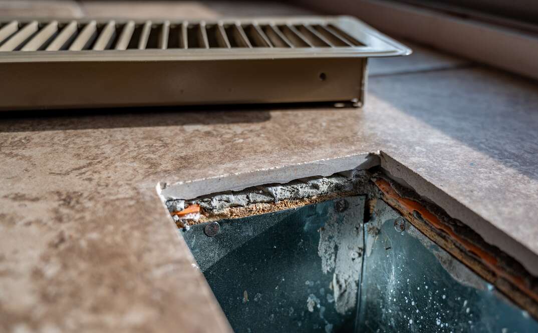 The grate for the HVAC return has been removed from the floor return and sits on the tile floor behind the open air duct beneath the flooring, air duct, duct, AC, air conditioning, heating, HVAC, heating ventilation and air conditioning, tile floor, tile, floor, AC return