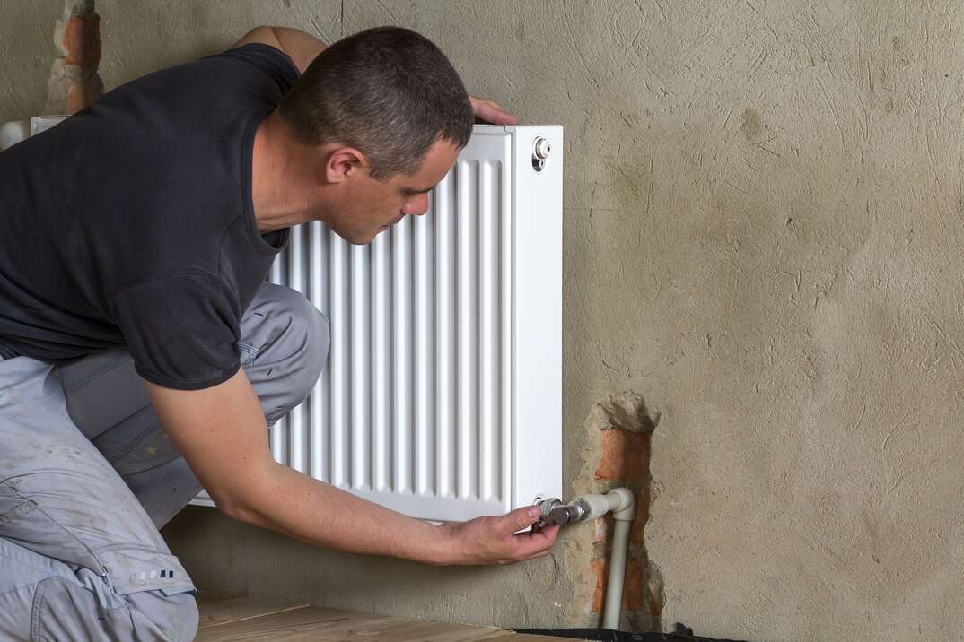 Young handsome professional plumber worker installing heating radiator on brick wall in an empty room of a newly built apartment or house. Construction, maintenance and repair concept.