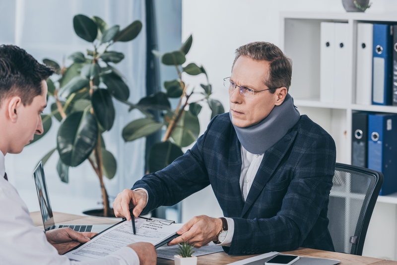 businessman in neck brace sitting at table and pointing on form for compensation claim to worker in office, compensation concept