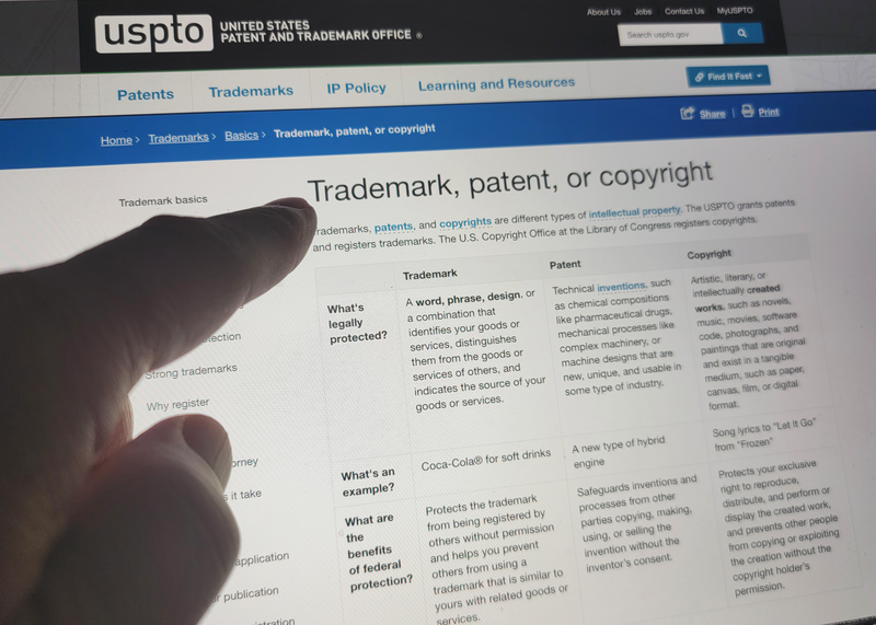 A human finger points to a glowing laptop computer screen displaying a government website defining the differences between copyright and patent and trademark, trademark, copyright, patent, website, computer, laptop