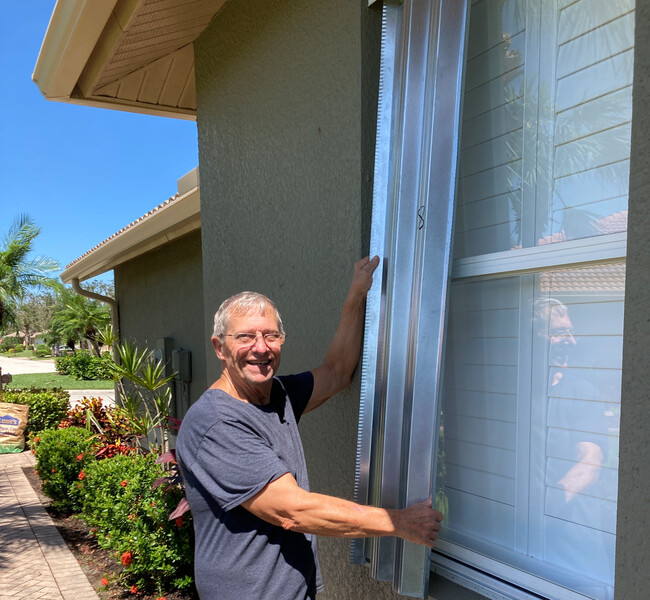 An older man installs hurricane shutters over an exterior window of a house with a blue sky and greenery in the background, hurricane, hurricane shutters, shutters, older man, blue sky, preparing for a storm, storm, weather