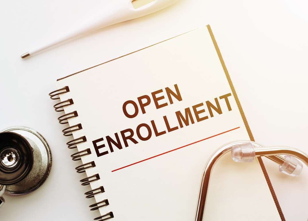 A health insurance open enrollment packet sits on a white table with a pen resting above it and a stethoscope resting on top of it, open enrollment packet, open enrollment, enrollment, health insurance, insurance, health, medical, paperwork, documents, stethoscope, white background, white table