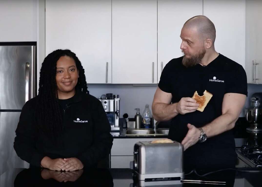 HomeServe Managing Editor Matt Schmitz eats a piece of toast at a kitchen counter while pushing the toaster over to HomeServe Editor Lauren Leazenby to offer her the other piece of toast, screen grab, video screen grab, thumbnail, video thumbnail, Lauren Leazenby, Matt Schmitz, toaster, toast, kitchen, kitchen counter, countertop, refrigerator, bread