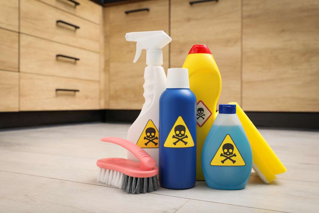 A grouping of different household cleaners with yellow triangular hazardous materials labels on them sits in on the white tile floor of a kitchen with light brown colored drawers and cabinetry in the background, hazardous materials, danger, skull and bones, crossbones, household cleaners, chemical cleaners, cleaners, cleaning solution, cleaning solutions, white tile floor, tile floor, tile, floor, kitchen floor, kitchen, kitchen cabinetry, cabinetry, light brown colored cabinetry, drawers, kitchen drawers, poison, danger, utility brush, brush, spray bottle, sprayer