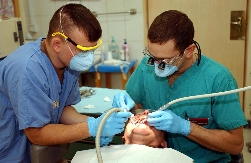 A male dentist and his assistant both wear medical scrubs and face masks and eyewear as they work on a patient in the dentist chair with a medical instrument in the dentist office, male dentist, dental hygienist, scrubs, dentist, dental, dentist's office, office, medical, medical office, dentist office, patient, dental patient, teeth, checkup, dental work, dental, medical, health, health care