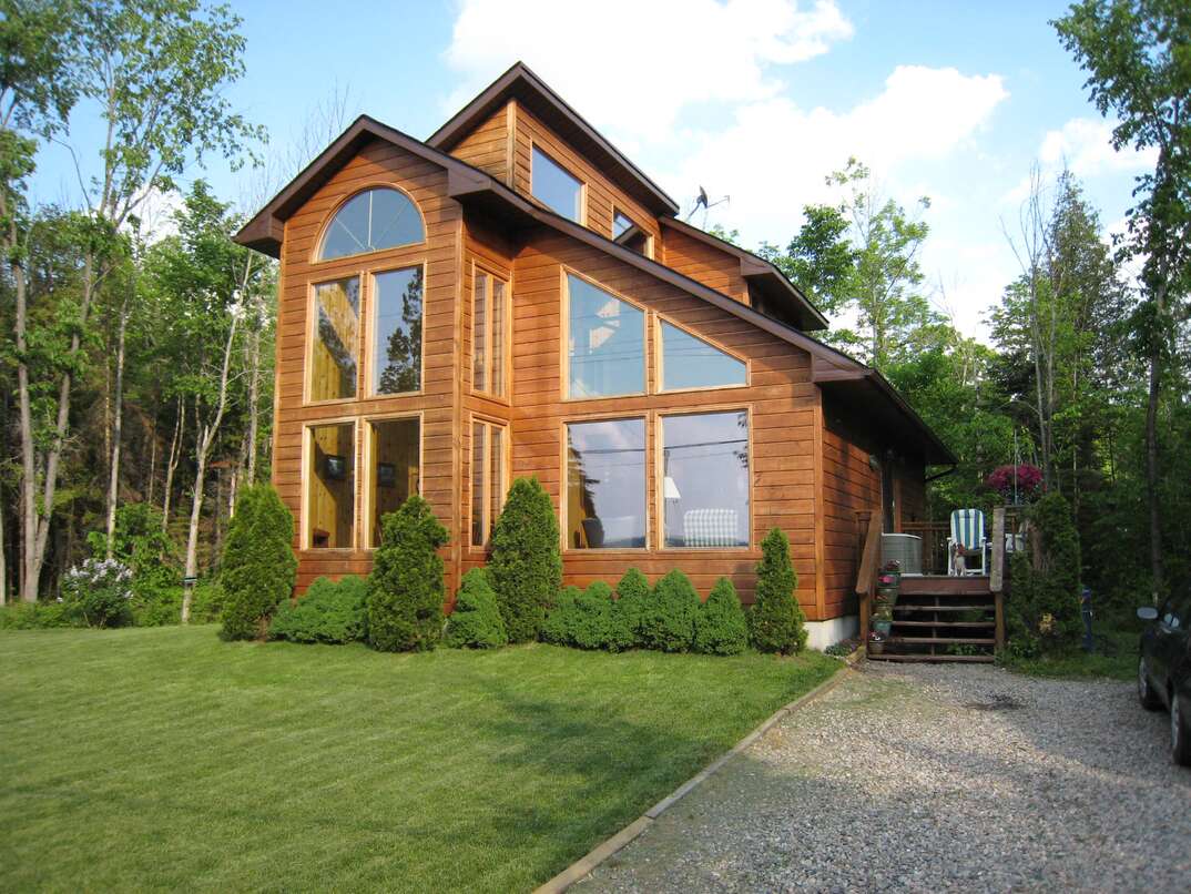 A house sits on a parcel of land with a green front lawn and surrounded by trees at the end of a gravel driveway, gravel driveway, gravel, driveway, house, brown house, wood house, wooden house, wooden, wood, trees, woods, forest, grass, green grass, lawn, green, green lawn, blue sky, white clouds, clouds