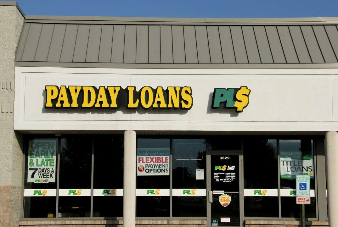 The storefront of a payday loans business is shown during the day in what appears to be a strip mall with no people in sight and a blue sky visible in the background above the roof of the building, payday loans, payday loan, payday, loan, predatory lending, lending, lender, bank, banking, finance, poor finances, strapped for cash, cash, money, strip mall, storefront, windows, blue sky