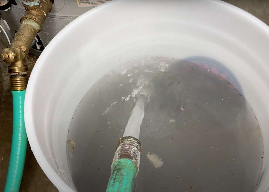 A hose empties into a bucket the dirty water and debris that comes out when you flush your water heater, water heater, hot water heater, dirty water, filthy water, dirty, filthy, hose, spraying, bucket, debris, maintenance, cleaning flushing, draining