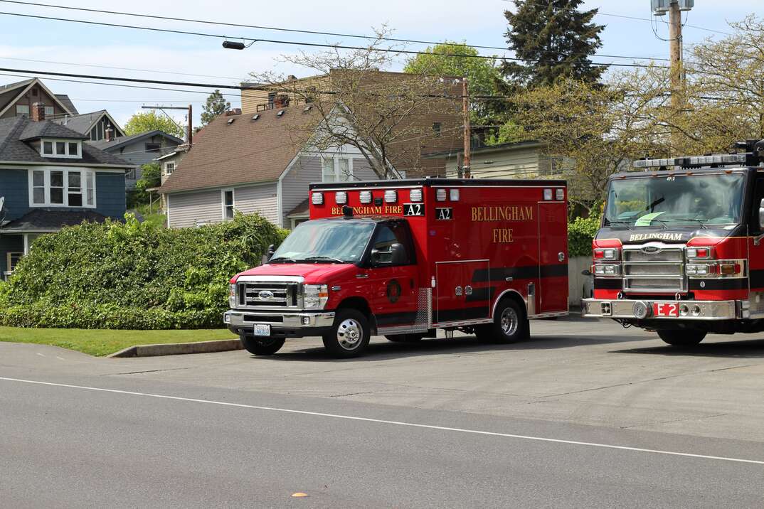 A red ambulance is stopped next to a red fire engine and ready to pull out onto a street with greenery and trees and houses in the background, ambulance, emergency services, emergency, firetruck, fire truck, fire engine, emergency vehicles, street, residential, residential street, houses, health, 911