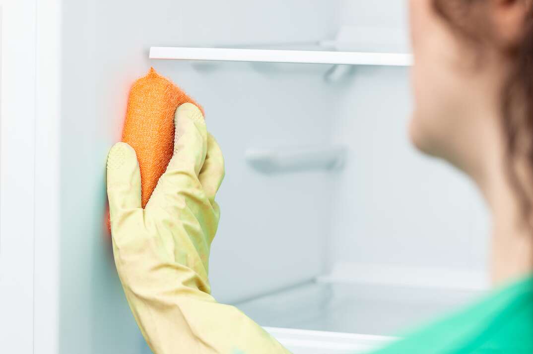A woman wearing a yellow latex glove scrubs the inside of a white freezer with an orange sponge