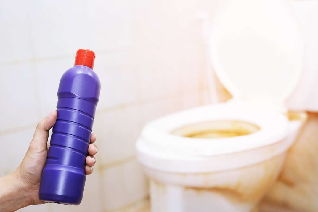 A human hand in the foreground holds a purple plastic bottle with a red cap against a white toilet with the seat up in a white bathroom, cleaner, bathroom cleaner, cleaning solution, cleaning, cleaner, toilet, toilet seat up, toilet seat, commode, bathroom, domestic bathroom, restroom, clean, cleaning product, purple bottle, bottle, plastic bottle, white bathroom, white tile, white toilet