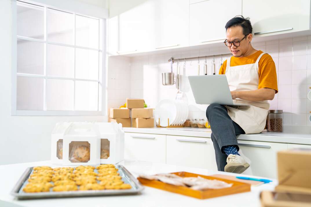 Portrait of Asian man bakery shop owner using laptop computer advertising online bakery store on social media in the kitchen, white kitchen, kitchen, man, male, baker, cookies, chocolate chip cookies, baking sheet, baking, baker, white apron, apron, laptop computer, laptop, computer, bakery, online bakery, home business, business, home kitchen, residential kitchen, online business, internet business, online, internet, cooking, cook, white countertop, countertop
