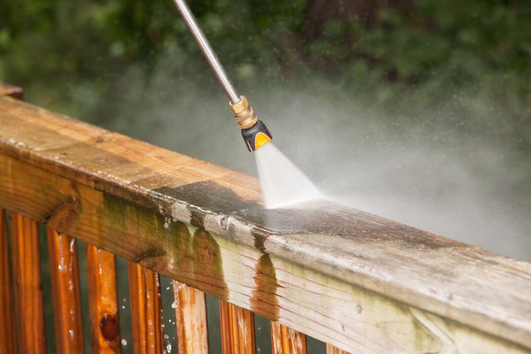 "A pressure washer sprayer is cleaning a weathered treated wood deck railing. The wood on the left has been  cleaned, the right is dirty and weathered, showing the contrast. The background are blurred trees."