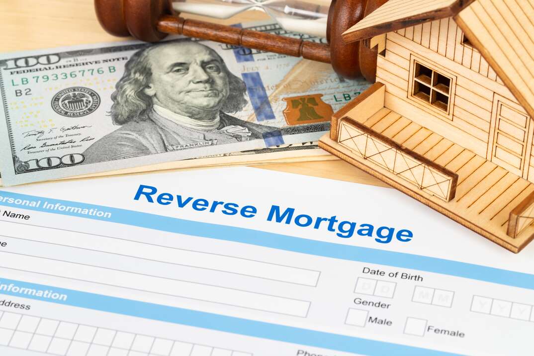 A reverse mortgage document sits on a table just below a stack of hundred dollar bills and a miniature model house, cash, money, hundred dollar bills, house, home, model house, miniature house, reverse mortgage, mortgage, home loan, form, document, legal document