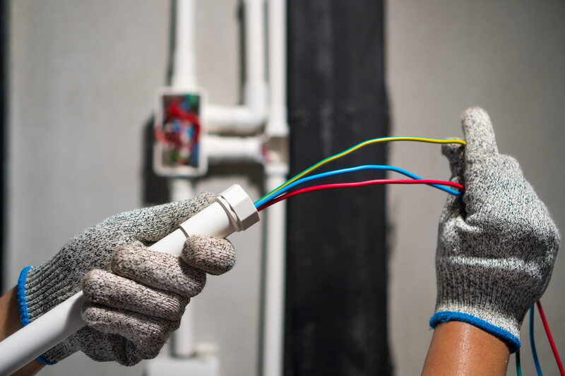 A closeup photo of the gloved hands of an electrician hold an electrical conduit with the left hand and several multicolored wires in the right hand against the backdrop of a gray wall with more conduits affixed to it, conduit, conduits, wiring, ground wire, wires, wire, electrician, electrical, electric, power, repair pro, technician, electrical technician, electric tech, gloves, gloved hands, hands
