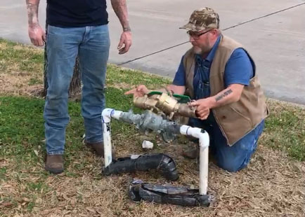A male plumbing contractor kneels next to a back flow preventer in a yard next to a street as another male contractor stands to the side looking on, plumbers, plumber, plumbing, contractors, maintenance workers, co-workers, men, males, yard, street, grass, green grass, backflow, preventer, backflow preventer, pipes, plumbing, exterior plumbing
