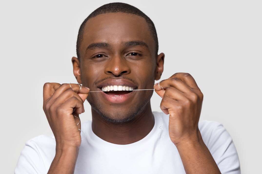 A man smiles as he holds a string of floss up to his teeth with both hands against a white background, man, man flossing, flossing, male, male flossing, dental floss, dental hygiene, oral, oral hygiene, white teeth, teeth, tooth, white background, white T-shirt, smiling, smile, oral health, dental health, tooth care