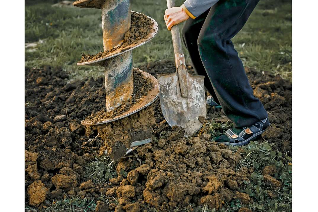 A pile of dirt is shown forming in a grassy yard as a construction auger drills as a man digs for the ostensible purpose of putting in a well, auger, drill, dig, dirt, mud, soil, grass, lawn, yard, green grass, brown mud, brown dirt, construction, construction equipment, person, man, dig, shovel, digging
