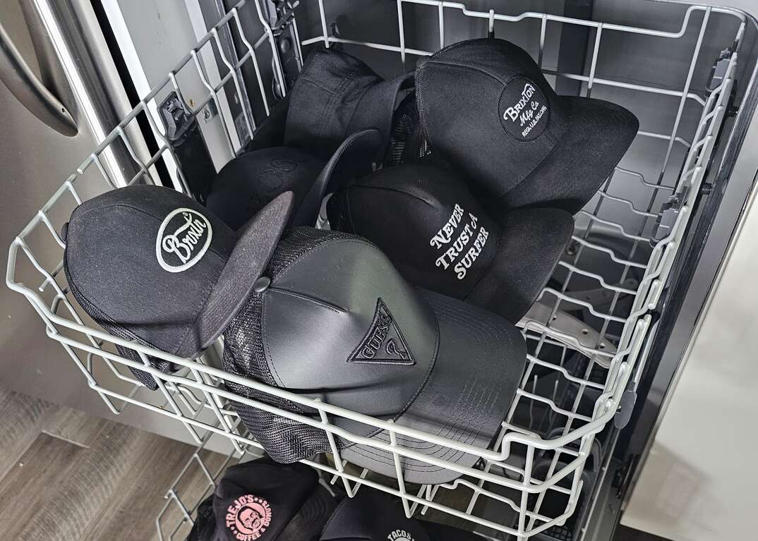 A dishwasher in a domestic kitchen has the door open and both the top and bottom racks pulled out and filled with baseball caps with varied logos on them, dishwasher, kitchen appliances, kitchen appliance, kitchen, modern kitchen, domestic kitchen, appliances, appliance, baseball caps, caps, hats, men's hats, headwear, gray hardwood floor, hardwood floor, floor, hardwood, kitchen floor, washing hats in dishwasher, washing hats, hats in dishwasher, dishwasher rack, rack, racks, top rack, bottom rack, open dishwasher