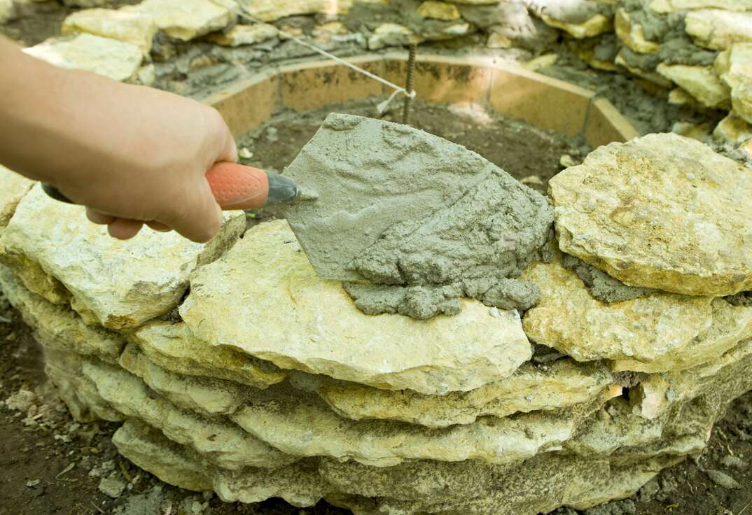 DIY homeowner Laying Mortar with a Trowel on Stones to create a backyard bonfire pit