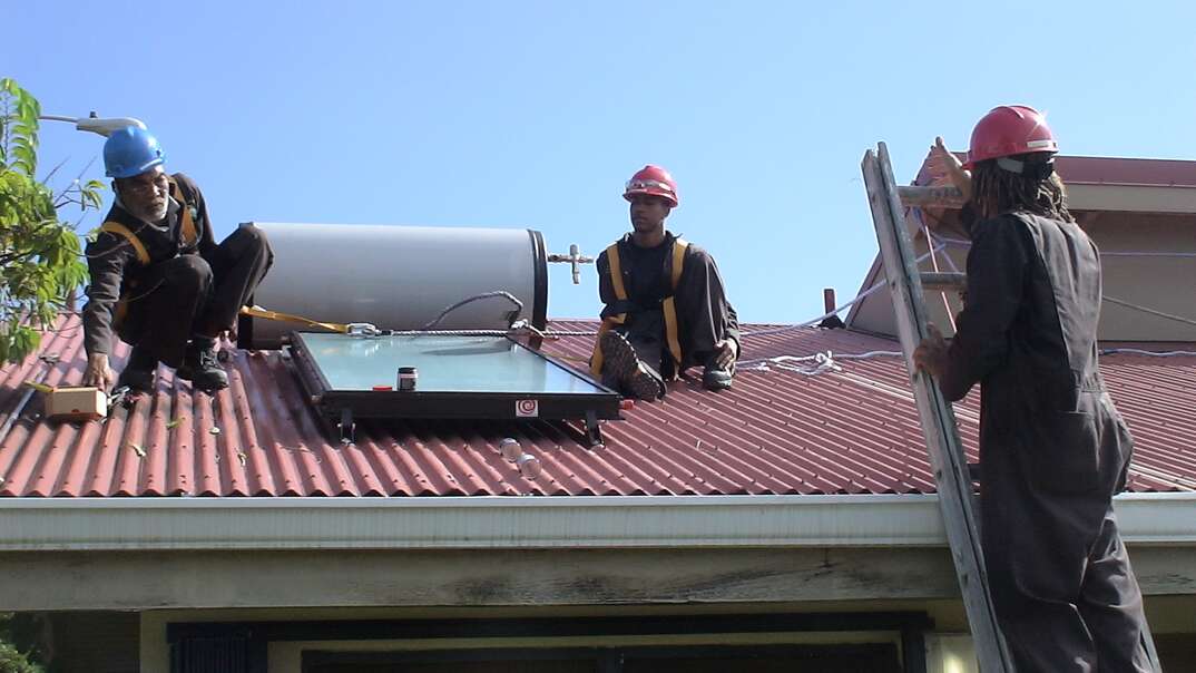 Two workers sit on a roof performing a solar water heater installation as a third worker reaches the top of a ladder leaning against the top of the house, workers, solar water heater, solar, water heater, water, heater, solar energy, solar power, solar heat, blue sky, sky, red roof, roof, contractors, ladder, man on ladder, house