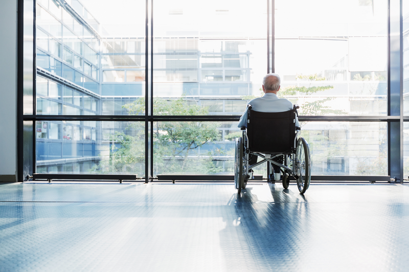 An elderly man sits in a wheelchair with his back to the viewer as he stares out the window of an institutional room into the courtyard of a modern building complex, elderly man, man, old man, wheelchair, nursing home, building complex, large glass window, window, elderly, old, mature, courtyard, sunny day