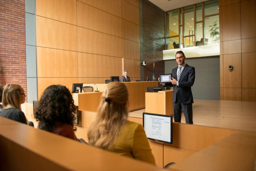A lawyer standing in the middle of a high ceiling courtroom addresses a group of jurors in the jury box as the judge looks on from the stand in the background, jury box, jury, trial, criminal trial, legal trial, case, court case, legal case, courtroom, judge, lawyer, attorney