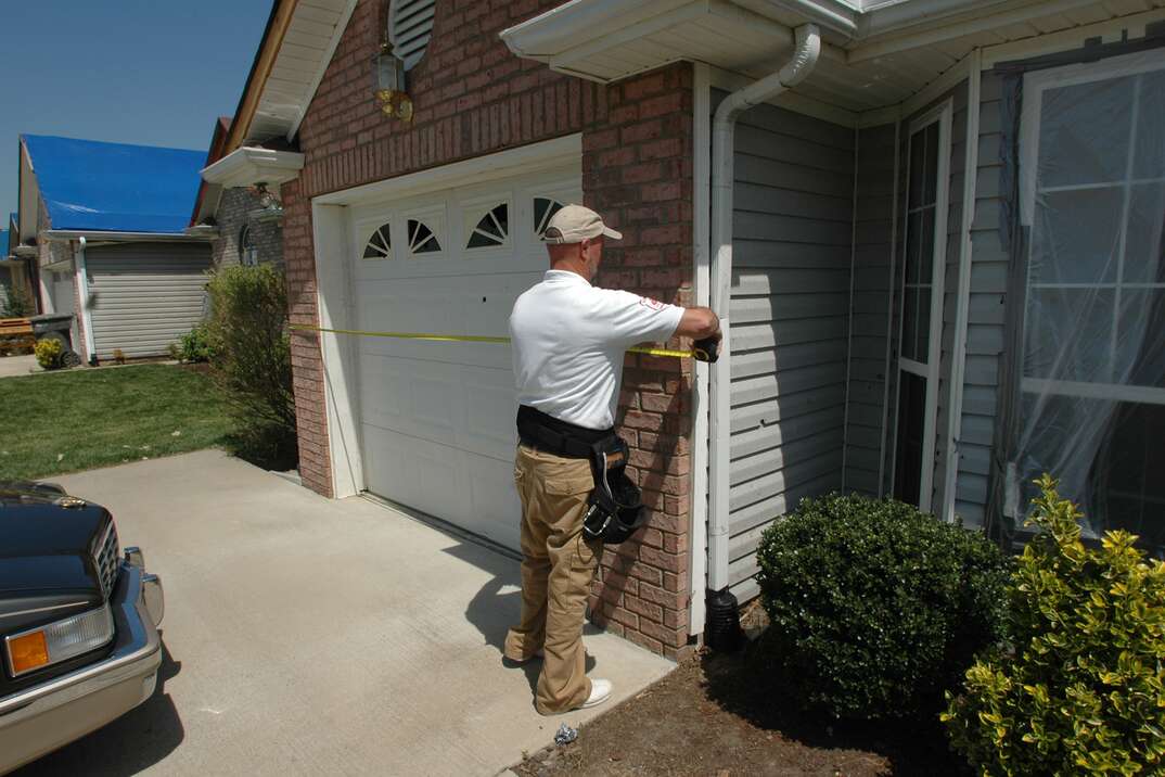 A male insurance adjustor stands in the driveway of a residential house as he inspects the building for damage following a tornado, tornado damage, damage, building, house, home inspector, inspector, driveway, brick house, brick, insurance, insurance adjustor, insurance adjuster, shrubbery