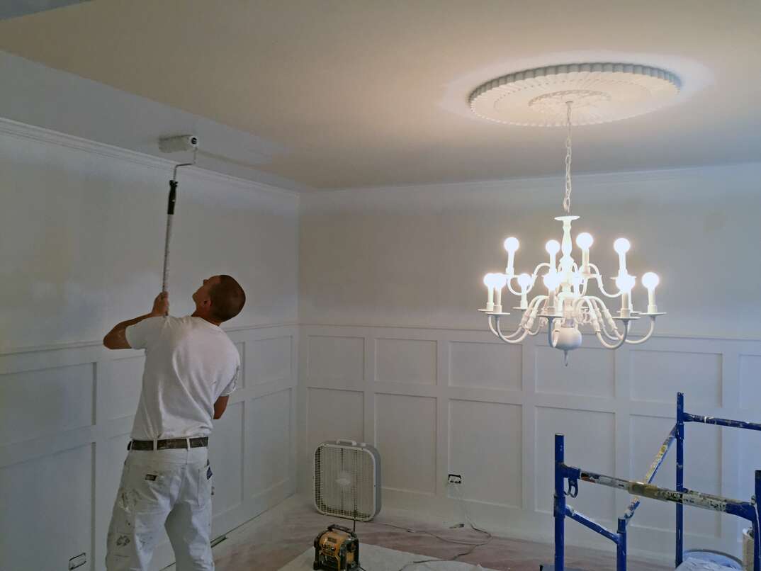 male painter wearing all white paints a ceiling white using a paint roller on a pole 