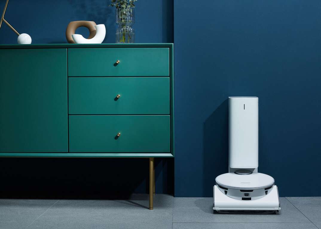 Samsung JetBot 90 AI Plus smart mop docked next to a green dresser with multiple drawers and knickknacks in a room with blue walls.