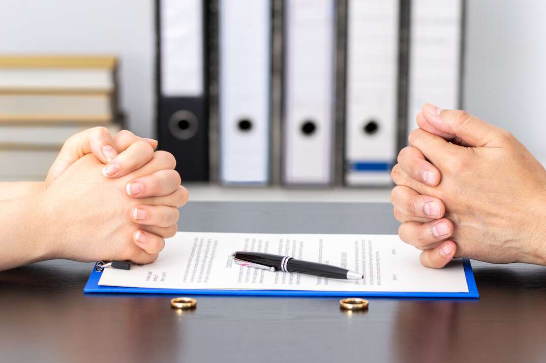 A pair of male hands and a pair of female hands rest on either side of a divorce document with a pen on top of it and gold rings beside it on the table, hands, male and female hands, couple, husband and wife, divorce document, divorce, document, legal document, legal, lawyer, lawyer's office, gold rings, rings, wedding rings, engagement rings, pen, signing document