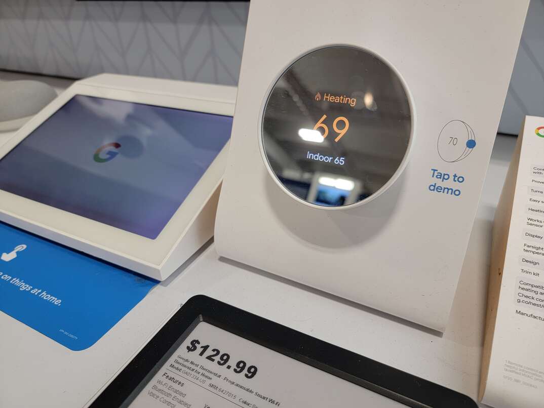 A Google Nest smart thermostat reading a temperature of 69 degrees on its round digital display sits in front of a price tag that reads  129.99, and next to a digital display screen showing a Google logo on it, both as part of an in-store retail display for shoppers.