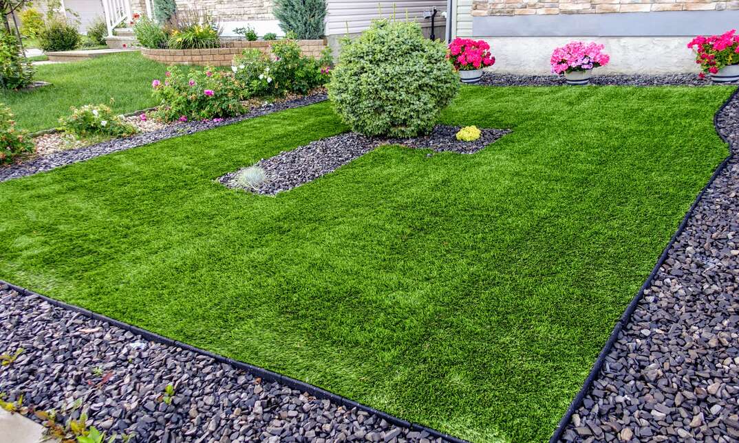 A green artificial turf lawn ish shown landscaped with a rectangular pebble border as well as green shrubbery and pink and red flowers, green artificial turf lawn, green artificial turf, artificial turf, turf, green, verdant, grass, lawn, yard, front yard, back yard, frontyard, backyard, shrubbery, shrubs, green shrubs, shrub, pebble border, pebbles, landscaping, landscaped, flowers, pink flowers, red flowers